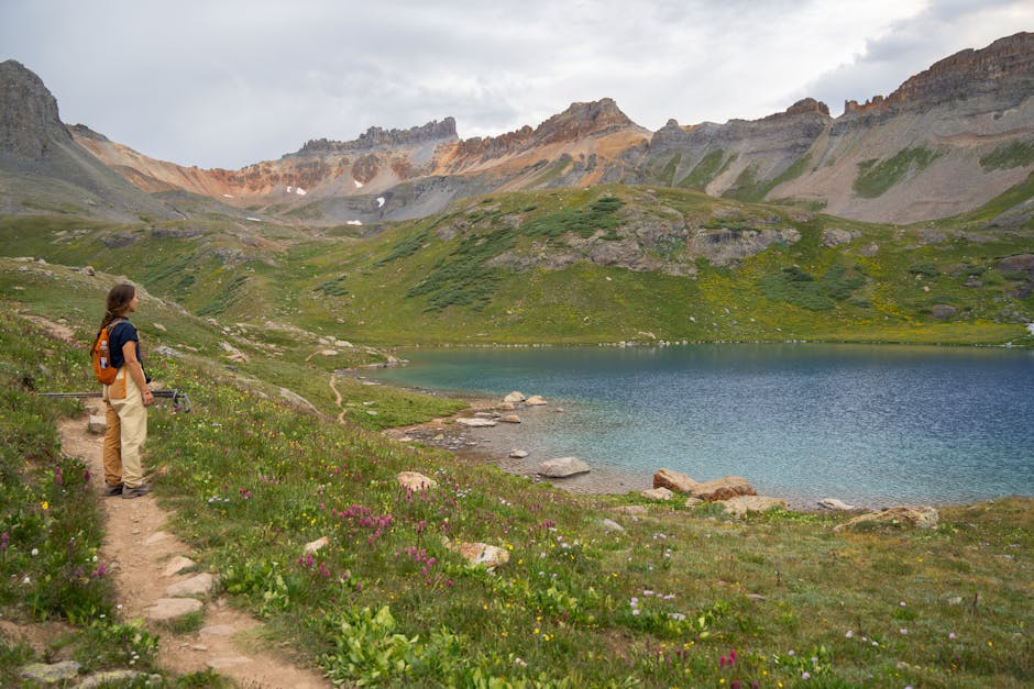 A woman hikes along a mountain trail by a serene blue lake surrounded by lush greenery.
